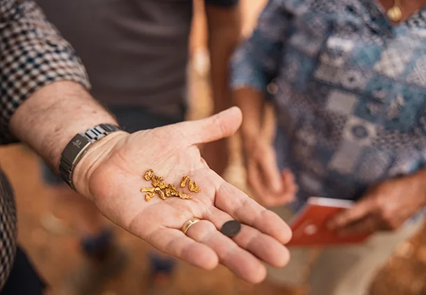 Close up of man's hand holding a gold nugget with blurred person in the background.